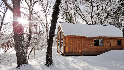 Chalet en bois recouvert de neige dans une forêt d’hiver, soleil à travers les arbres, Huttopia Winter Chalets.