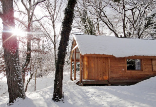 Cabaña de madera cubierta de nieve en un bosque invernal, luz solar entre los árboles, Huttopia Winter Chalets.