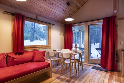 Acogedor chalet de madera con cortinas rojas y sofá, en Huttopia, con vistas al bosque cubierto de nieve.