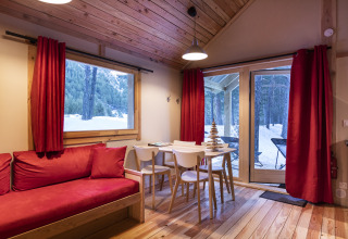 Acogedor chalet de madera con cortinas rojas y sofá, en Huttopia, con vistas al bosque cubierto de nieve.