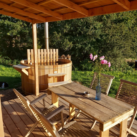 Terraza de madera en Dihan Evasion, Bretaña, con jacuzzi, mesa, sillas y flores, rodeada de naturaleza.