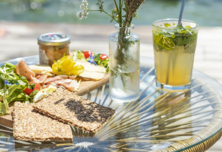 Buitenlunch met knäckebröd, ham, kaas, verse salade en een frisse drank op een glazen tafel.