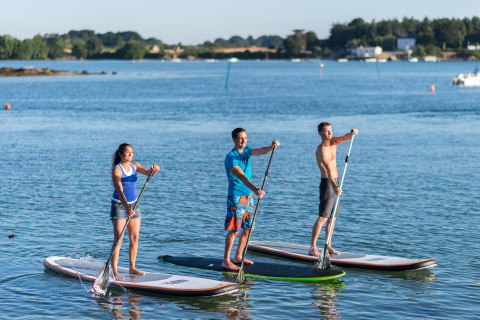 Tre persone fanno paddleboard in piedi su un lago presso Dihan Evasion - Lodges Bretagne, con il sole.