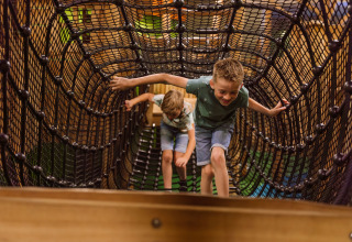 Two children play on a rope tunnel in the indoor playground at Vakantiepark Ommerland – Safaritenten Overijssel.