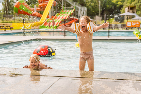 Two kids play and splash water in the children’s pool at Vakantiepark Ommerland with colorful slides visible.