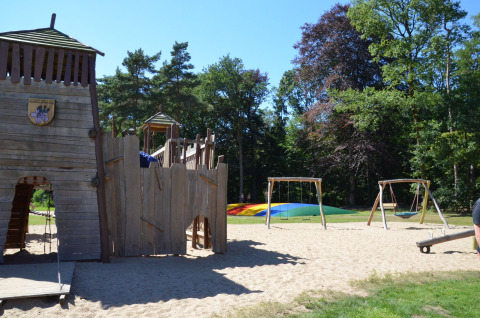 Aire de jeux pour enfants avec château en bois, balançoires et coussin sauteur coloré à Vakantiepark Ommerland.