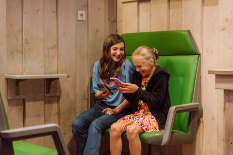 Due bambini felici seduti su sedie verdi all'interno di Vakantiepark Ommerland – Safaritenten Overijssel usano i telefoni.