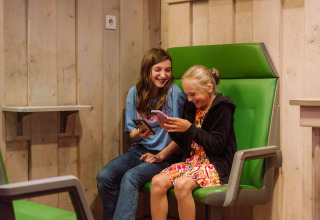 Zwei fröhliche Kinder sitzen drinnen auf grünen Stühlen im Vakantiepark Ommerland – Safaritenten Overijssel und nutzen Handys.