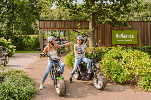 Two women on e-scooters in front of a sign reading 'NuNature outdoor + events' surrounded by nature.