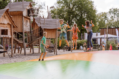 Kinder springen auf einem Hüpfkissen vor Holzhütten im Vakantiepark Ommerland – Safaritenten Overijssel.