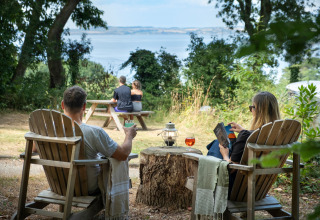 Twee stellen ontspannen bij Huttopia Douarnenez – Glamping Bretagne met uitzicht op bos en zee vanuit houten stoelen.