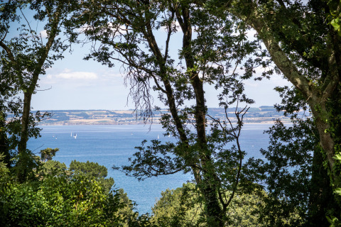 Vista al mar entre árboles en Huttopia Douarnenez – Glamping Bretagne, con veleros a lo lejos.