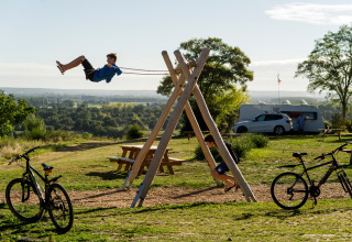 Børn leger på en gynge med cykler parkeret foran og campingvogne i baggrunden ved Huttopia Saumur.