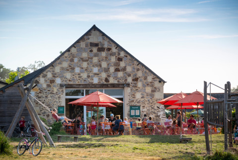 Café al aire libre con sombrillas rojas, huéspedes y parque infantil en Huttopia Saumur - Glamping Pays de la Loire.