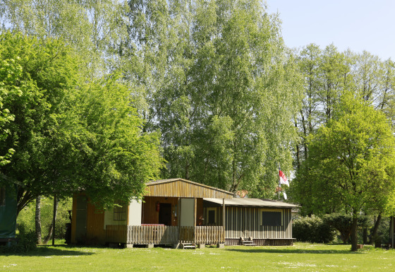 Glamping-Unterkunft bei Kano & Kamp Aqua Hema- Blokhutten Beieren, umgeben von grünen Bäumen und Sonne.