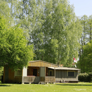 Glamping indkvartering på Kano & Kamp Aqua Hema- Blokhutten Beieren, omgivet af grønne træer og sollys.