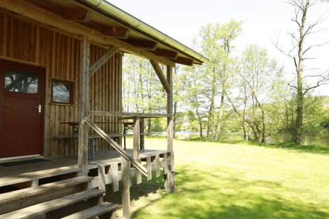 Wooden cabin at Kano & Kamp Aqua Hema- Blokhutten Beieren with porch, grassy area, and lakeside view.