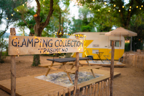 Cartel que dice 'Glamping Collection Trasimeno', con una caravana y luces colgantes en un entorno natural acogedor.