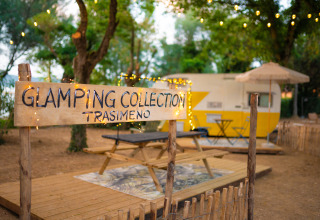 Cartel que dice 'Glamping Collection Trasimeno', con una caravana y luces colgantes en un entorno natural acogedor.