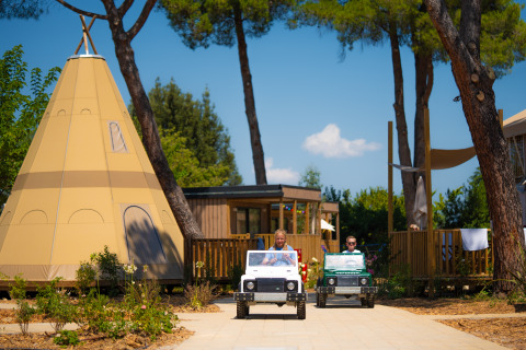 Bambini guidano mini jeep davanti a una tenda tipi e lodge al Trasimeno Glamping Resort in Italia.