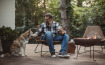 Hombre relajándose con café y su perro junto a una fogata en la terraza de un parque de glamping.