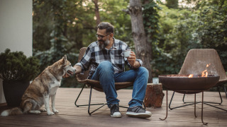 Hombre relajándose con café y su perro junto a una fogata en la terraza de un parque de glamping.