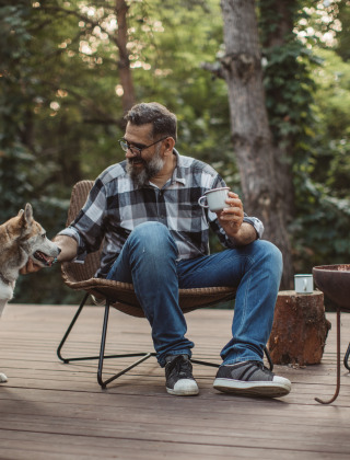 Mand slapper af med kaffe og hund ved bålsted på en terrasse i en grøn, skovklædt glamping park.