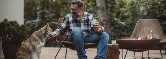 Hombre relajándose con café y su perro junto a una fogata en la terraza de un parque de glamping.
