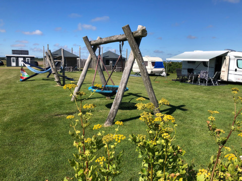 Camping Duinzoomhoeve glamping med legehus, gynger, campingvogne og farverige blomster i solrige omgivelser.