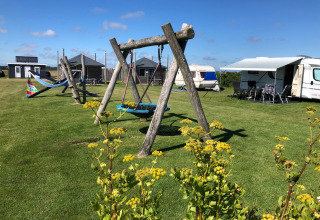 Camping Duinzoomhoeve glamping med legehus, gynger, campingvogne og farverige blomster i solrige omgivelser.