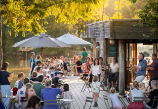 People relaxing outdoors by a lake café with live saxophone music at Village Huttopia Senonches - Glamping Val de Loire.