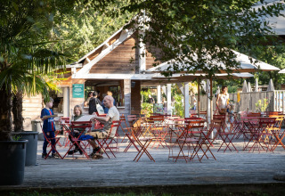 Area esterna a Huttopia Beaulieu sur Dordogne con famiglie, bambini, alberi e tavoli rossi all’ombra.