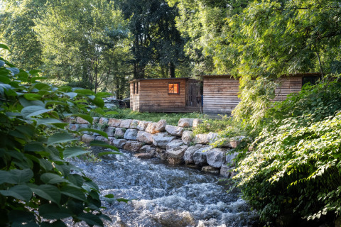 Wooden cabins by a flowing river at Huttopia Beaulieu sur Dordogne – Glamping Dordogne, surrounded by trees.