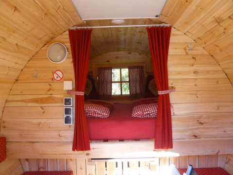 Interior de un barril de glamping en Baden-Württemberg, con paneles de madera, cortinas rojas y cama acogedora.
