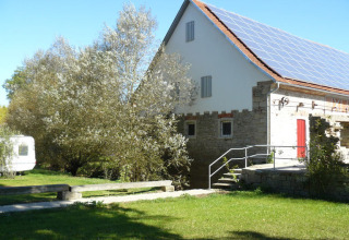 Día soleado en Camping Schwabenmühle, paneles solares en el techo, árboles verdes y caravana blanca.