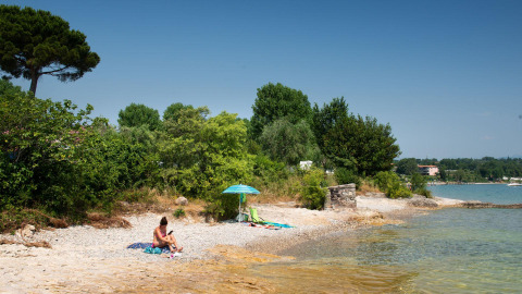 Vrouw ontspant zich bij Sivinos Camping Boutique aan het Gardameer op een kiezelstrand met bomen en zon.