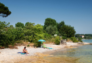 Vrouw ontspant zich bij Sivinos Camping Boutique aan het Gardameer op een kiezelstrand met bomen en zon.