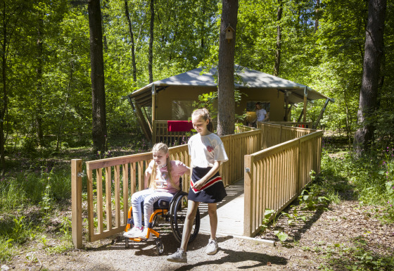 Zwei Kinder, eines im Rollstuhl, auf einer Rampe vor einem barrierefreien Glampingzelt im Wald.
