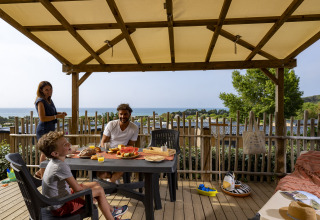 Famiglia fa colazione su terrazza in legno con vista mare al glamping Camping Saint Pabu Plage in Bretagna.