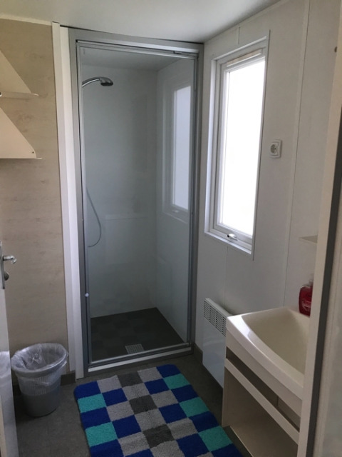 Moderne badkamer met douchecabine, wastafel, raam en kleurrijk geblokt vloerkleed in glamping accommodatie.