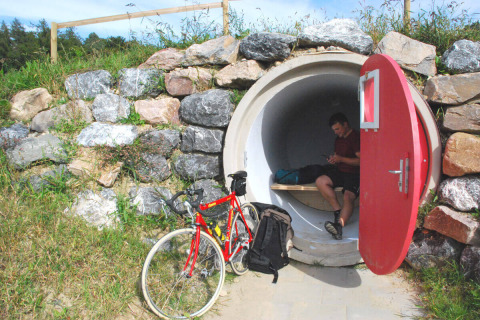 Cykelist hviler sig i et rørformet glamping-hus med rød dør, omgivet af sten og græs hos Camping & Vakantiepark Orsingen.