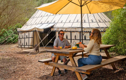 Dos personas disfrutan de un desayuno en una mesa de picnic bajo una sombrilla frente a una tienda glamping en Refugio Marnes.