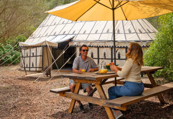Two people enjoy breakfast at a picnic table under a parasol in front of a glamping tent at Refugio Marnes.