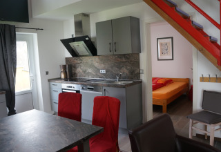 Modern kitchen and dining area with red chairs, staircase, and bedroom in Camping Altschmiede lodge.