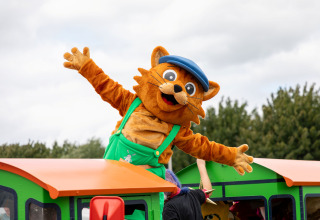Maskottchen im Katzenkostüm mit blauer Kappe winkt auf Spielzug im Safarizelt Gelderland Campingplatz.