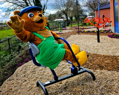 Mascotte in groene overall en blauwe pet zwaait op speeltuin bij Vakantiepark De Oude Molen - Safaritent Gelderland.