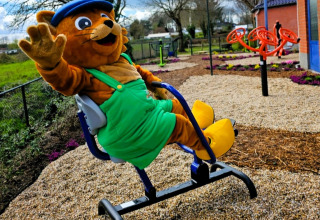 Mascot in green overalls and blue cap waves on playground at Vakantiepark De Oude Molen - Safaritent Gelderland.