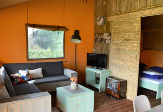 Interior de alojamiento glamping con sofá, TV, muebles de madera y dormitorio en tienda safari en Gelderland.