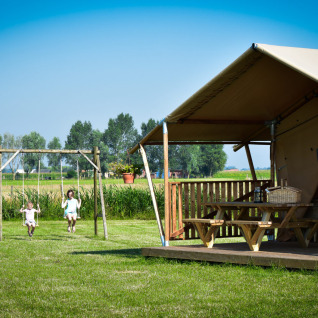 Glamping - aan de Vleterbeke: Børn leger på gynger nær luksustelt på grøn mark under en blå himmel.