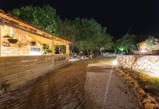 Vue nocturne de Camping Nono Ban - Safaritenten Brac avec allées éclairées, arbres et bâtiments en bois rustiques.